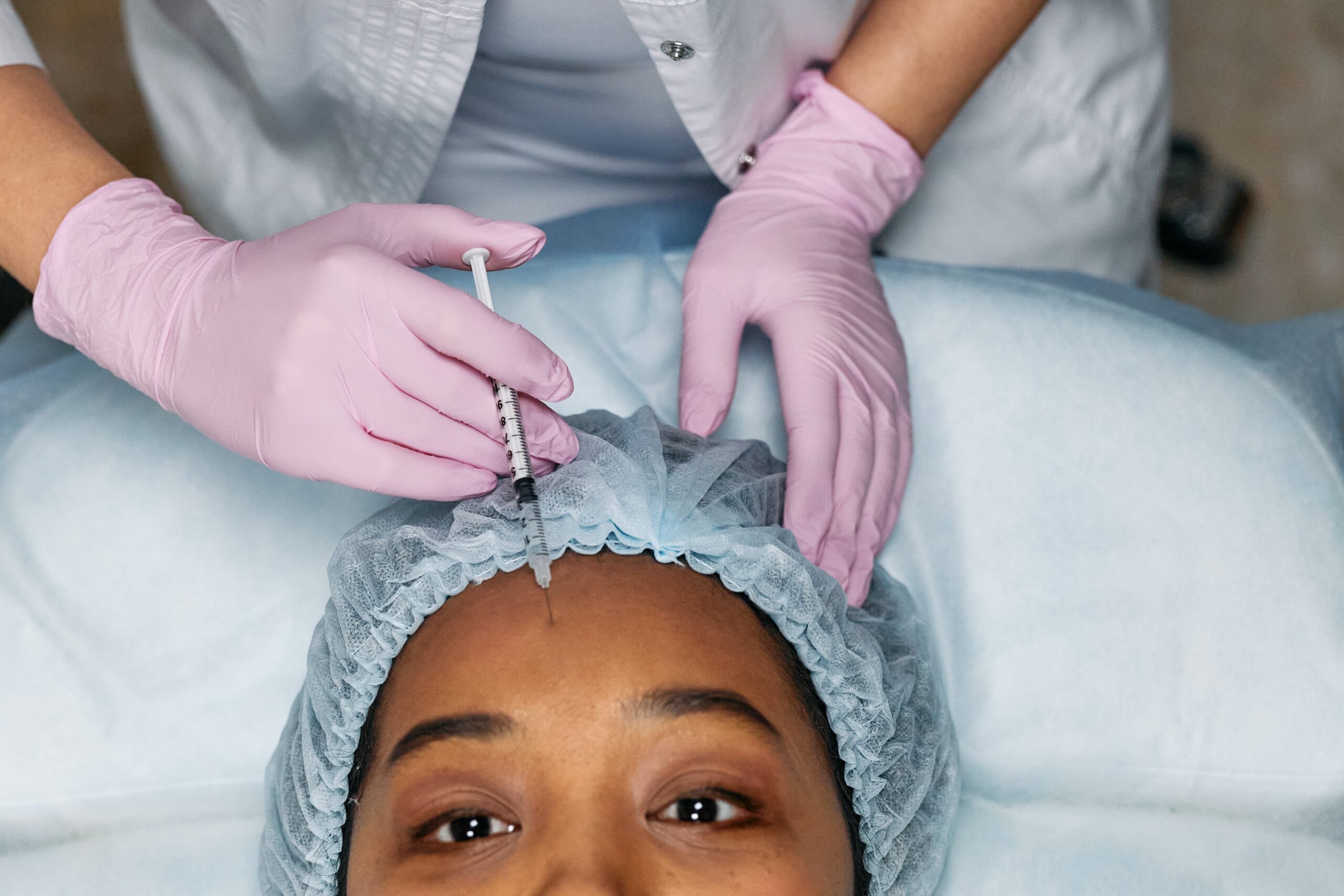 Picture of somebody getting anti-wrinkle injections or botox in their forehead, performed by an aesthetics practitioner with pink gloves.